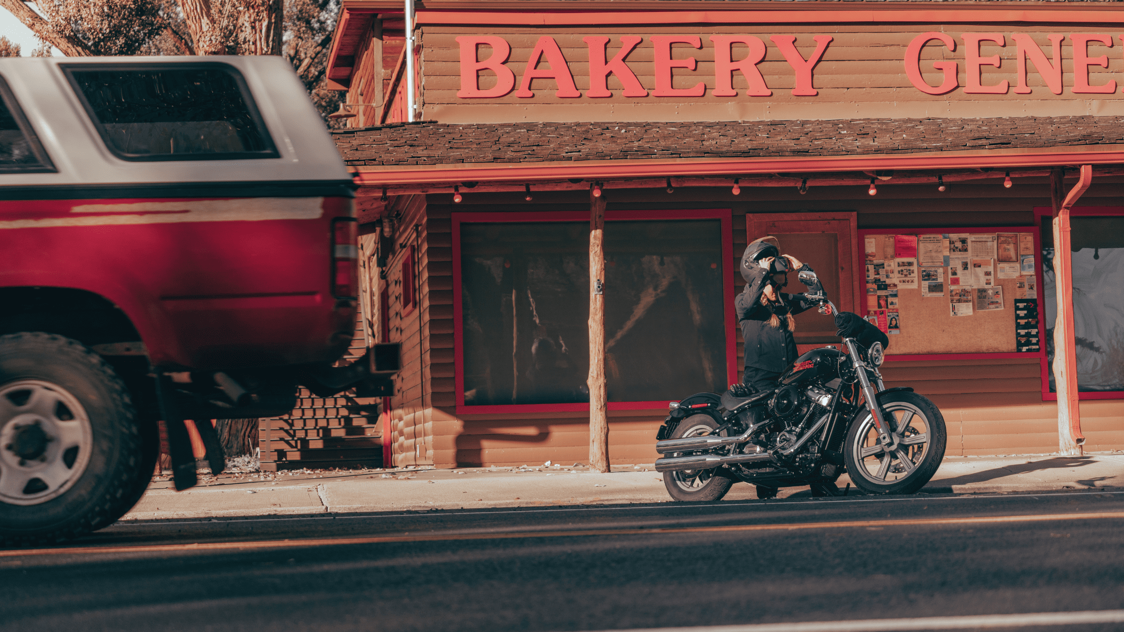 A person adjusts their helmet while standing beside a black motorcycle in front of a bakery store, with a red truck passing by.